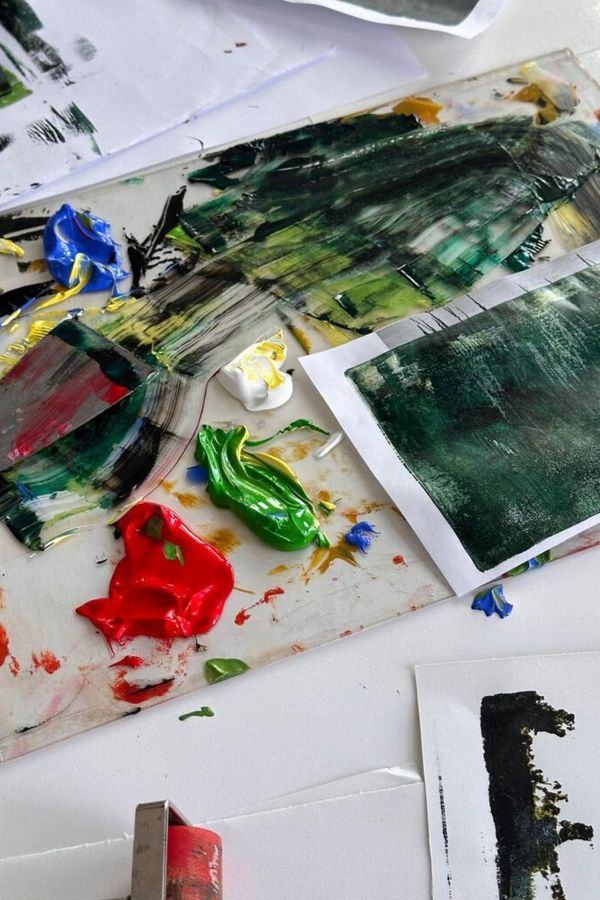 Czysta paleta z plamami zielonej, czerwonej, żółtej i niebieskiej farby olejnej, a obok niej szpachla malarska, wałek malarski oraz kartki papieru z fakturami farby.