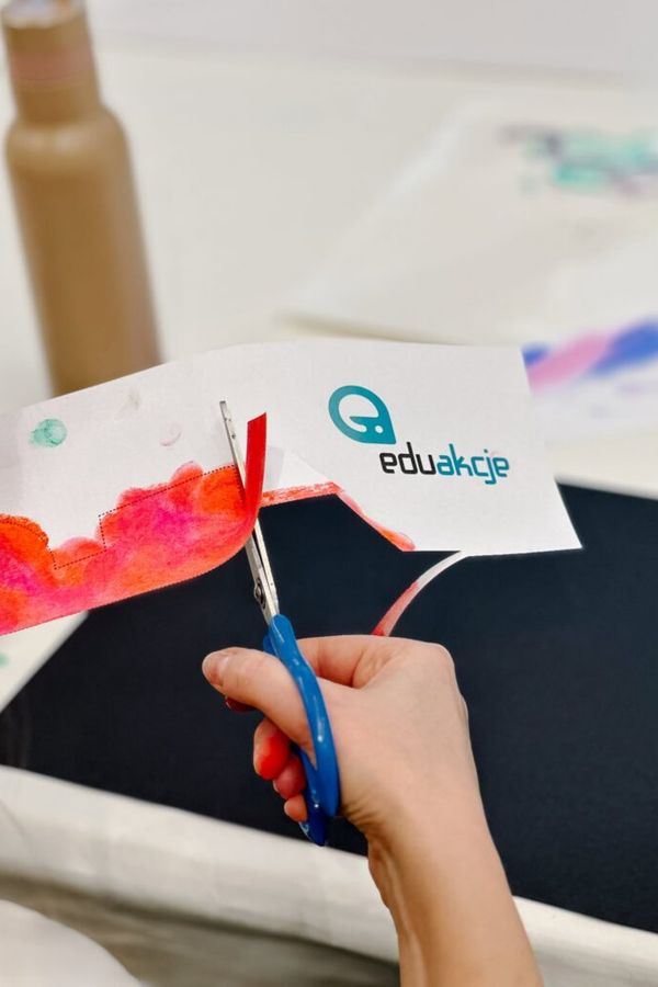 Ręka używa niebieskich nożyczek do przycięcia białego paska papieru z logo „eduakcje” i czerwonym wzorem namalowanym akwarelą.