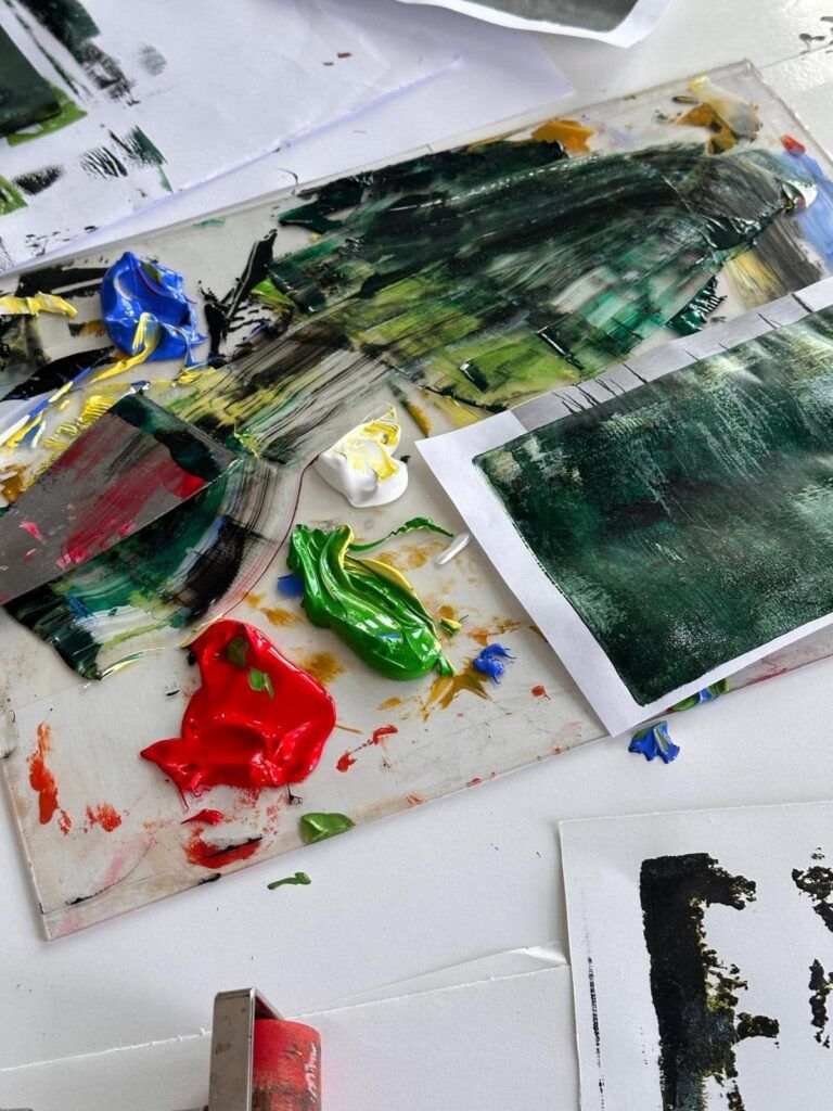 Czysta paleta z plamami zielonej, czerwonej, żółtej i niebieskiej farby olejnej, a obok niej szpachla malarska, wałek malarski oraz kartki papieru z fakturami farby.