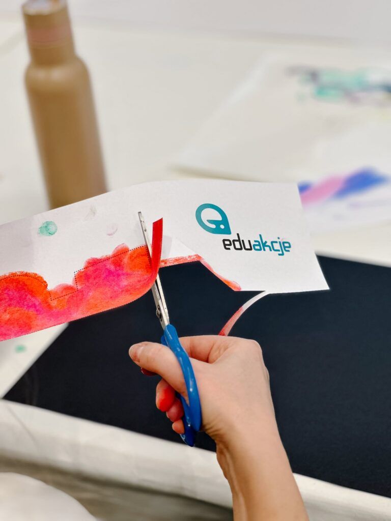 Ręka używa niebieskich nożyczek do przycięcia białego paska papieru z logo „eduakcje” i czerwonym wzorem namalowanym akwarelą.