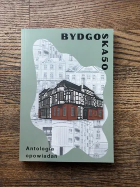 Na drewnianej powierzchni leży książka zatytułowana „BYDGO SKA50 Antologia opowiadań”. Na okładce widnieje ilustracja przedstawiająca budynek z muru pruskiego, a w tle szkice architektoniczne.