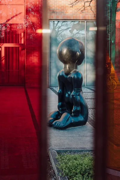 On the pavement near a modern glass building stands a large, dark sculpture depicting a kneeling figure resembling a cartoon character. The photograph was taken through a window with red and transparent panes.