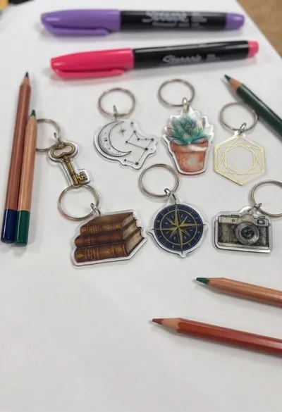 Na białej powierzchni ułożono sześć ilustrowanych breloczków, kredki oraz dwa markery Sharpie. Na breloczkach widnieją: klucz, księżyc, sukulent, kształt geometryczny, książki, kompas i aparat fotograficzny.