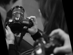 Czarno-białe zdjęcie przedstawiające dłonie trzymające aparat fotograficzny. Informacja o tytule i dacie wydarzenia.